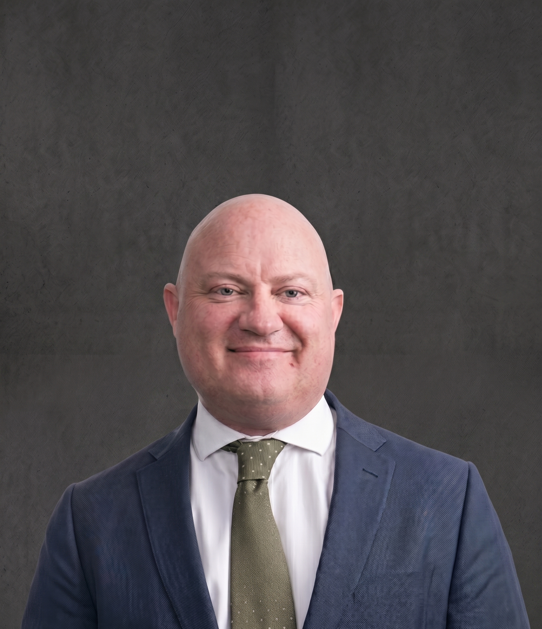 A smiling man in a navy suit, white shirt, and olive green tie against a dark gray textured background.