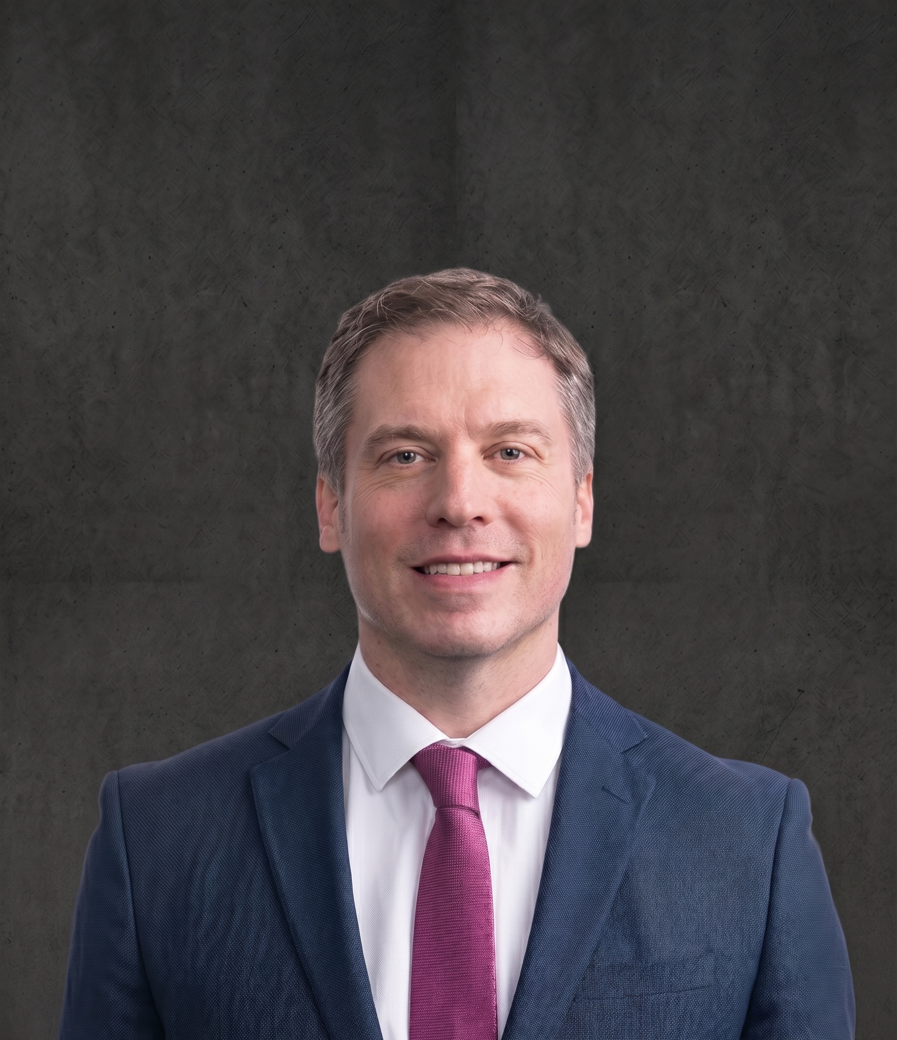 A man with gray hair and blue eyes wearing a dark blue suit jacket, a white dress shirt, and a pink tie, smiling in front of a dark background.
