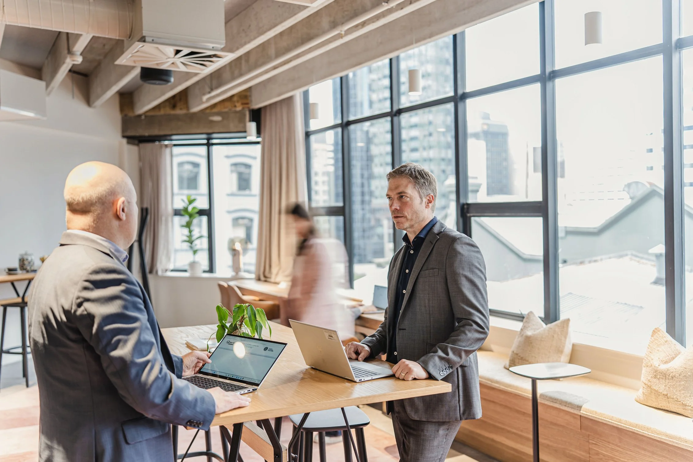 Two men in business attire having a discussion in a modern office space with large windows, laptops, and a plant on a wooden table.
