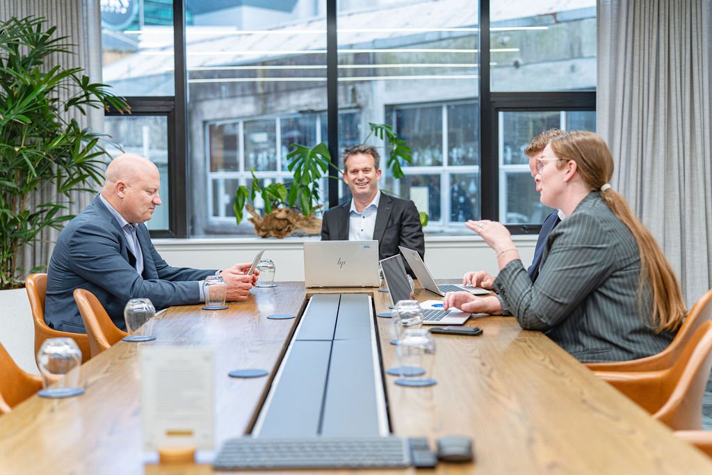 Five businesspeople in a meeting room having a discussion, with laptops and tablets on the table.