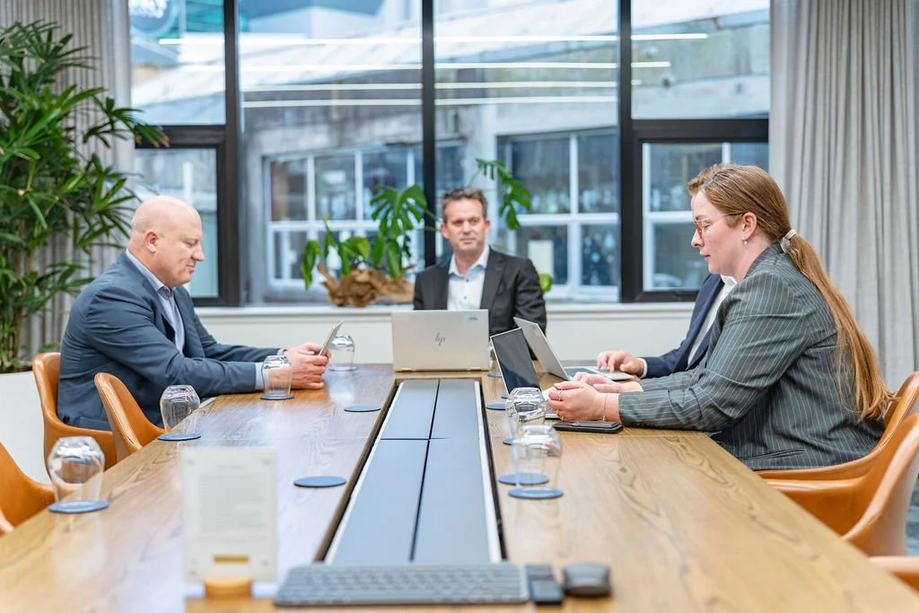 Five professionals in a meeting in a modern office conference room with large windows and plants.