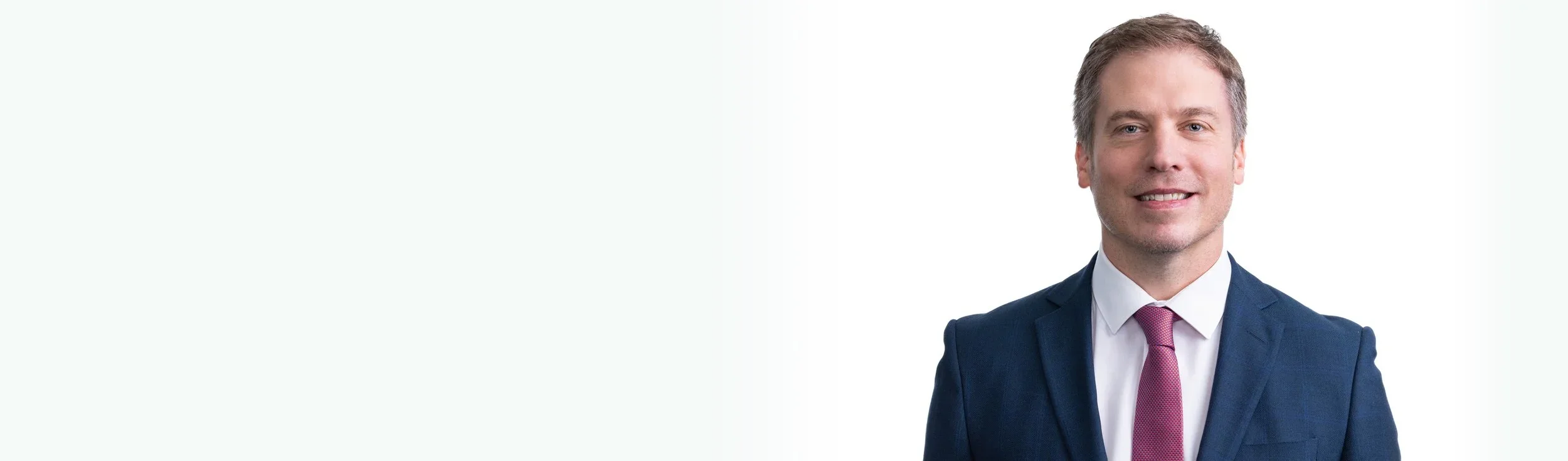 A smiling man in a dark blue suit with a white shirt and a pink tie, standing against a plain white background.