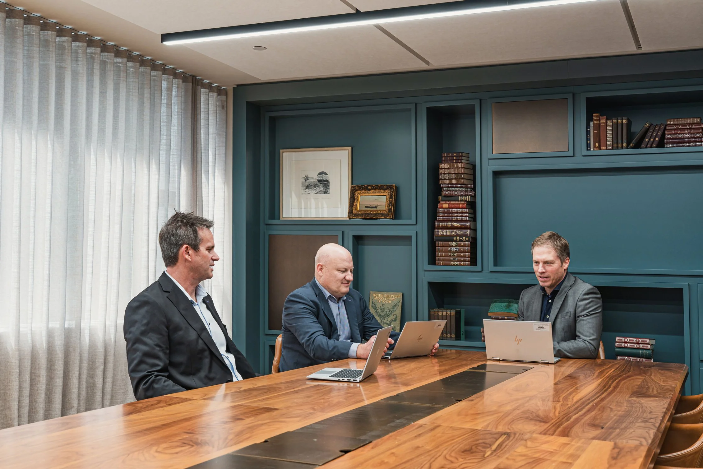 Three men in business attire sitting at a wooden conference table with laptops, engaged in discussion, in a room with teal wall-mounted bookshelves and framed artwork, curtains, and ceiling lights.