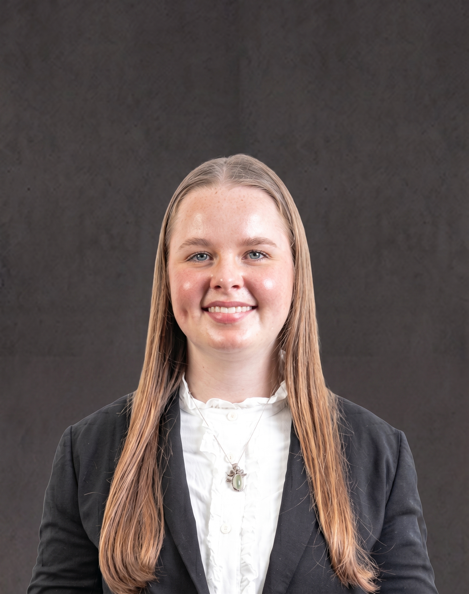 Young woman with long blonde hair, blue eyes, wearing a black blazer, white shirt, and a necklace with a large oval pendant, smiling against a dark background.