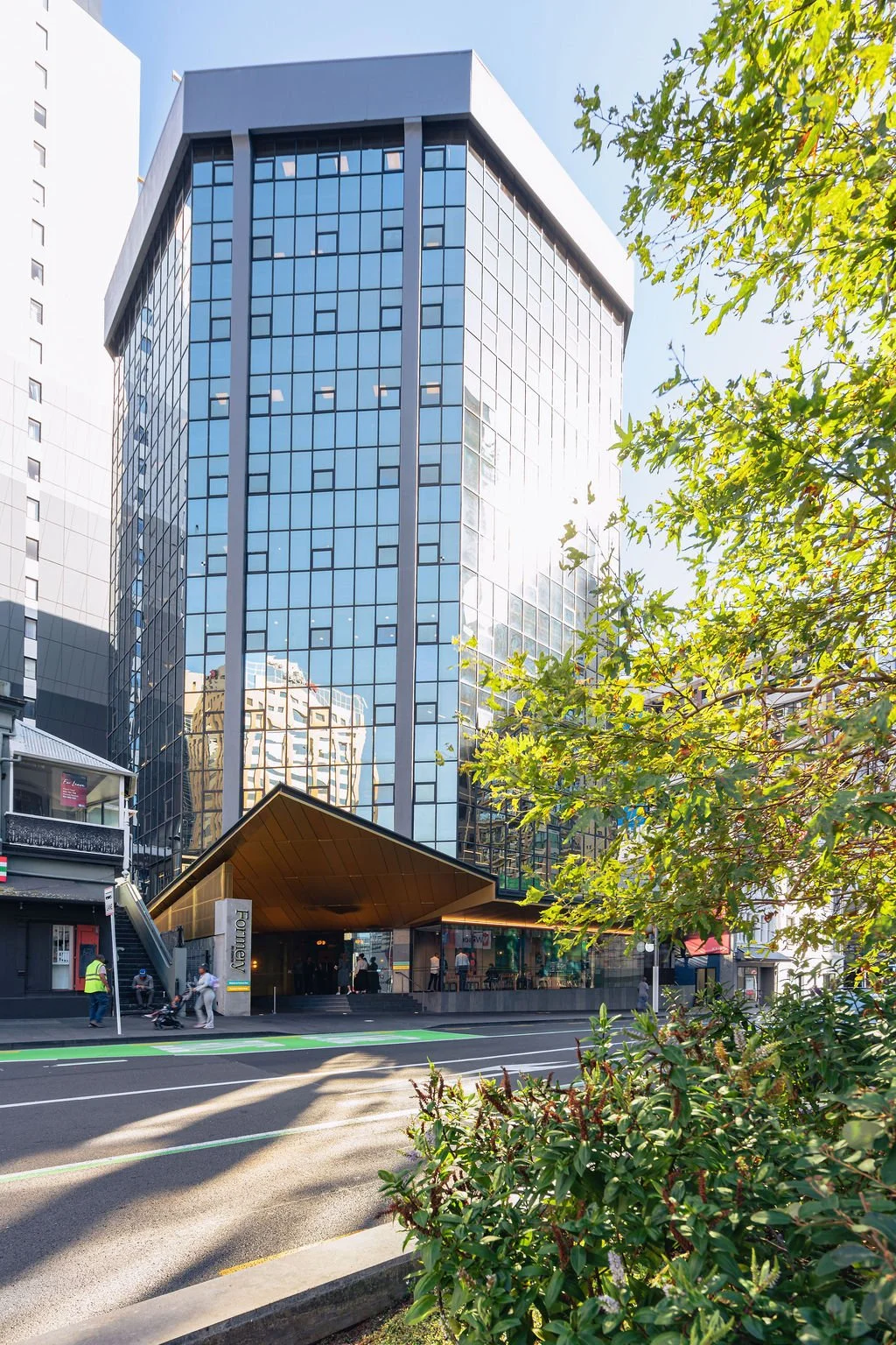 A modern glass office building with reflective windows, a gold-colored canopy entrance, and a sidewalk with pedestrians, trees with green leaves, and a street in front.