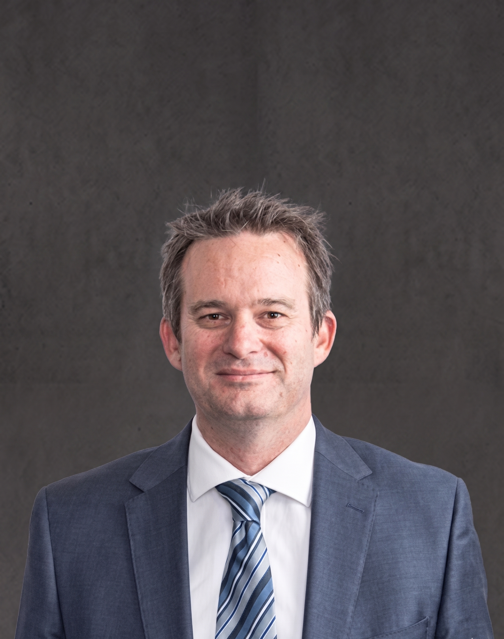 Close-up of a middle-aged man with gray hair, dressed in a dark suit, white shirt, and striped tie, against a dark gray background.