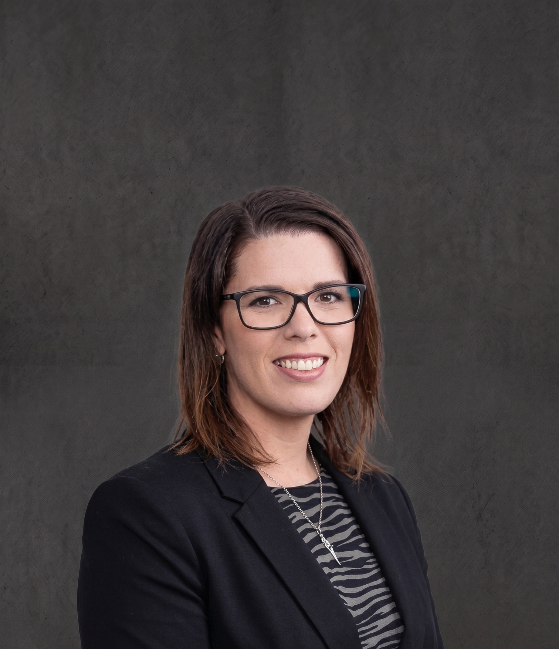 Close-up of a woman with brown hair, wearing glasses, a black blazer, and a striped shirt, smiling against a plain dark background.
