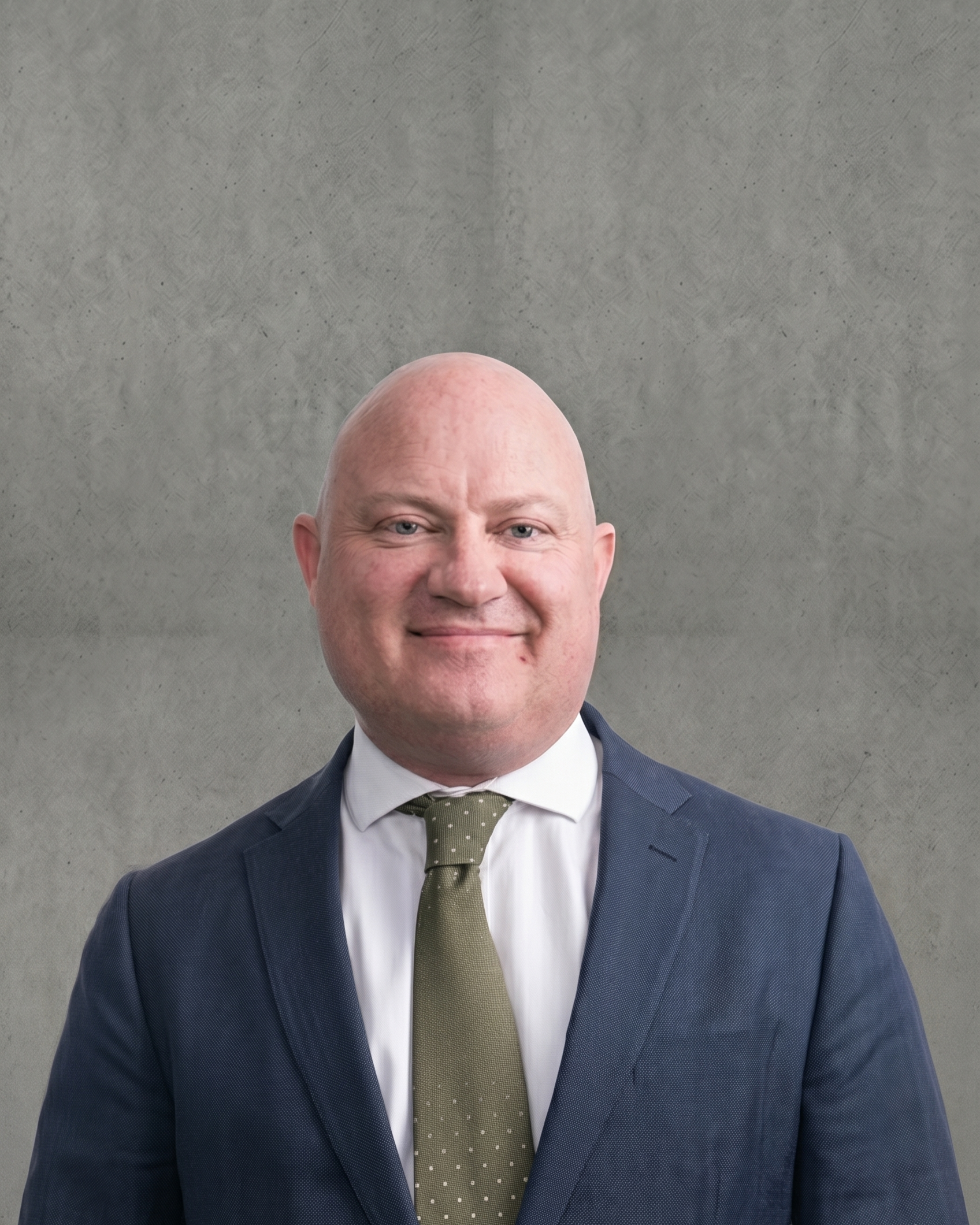 Close-up of a bald man in a navy blue suit, white shirt, and olive green tie with small white polka dots, smiling against a plain gray background.