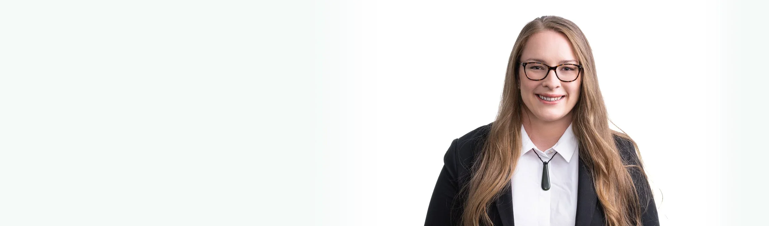 A young woman with long blonde hair, wearing glasses, a white shirt, a black blazer, and a black pendant necklace, smiling at the camera against a plain white background.