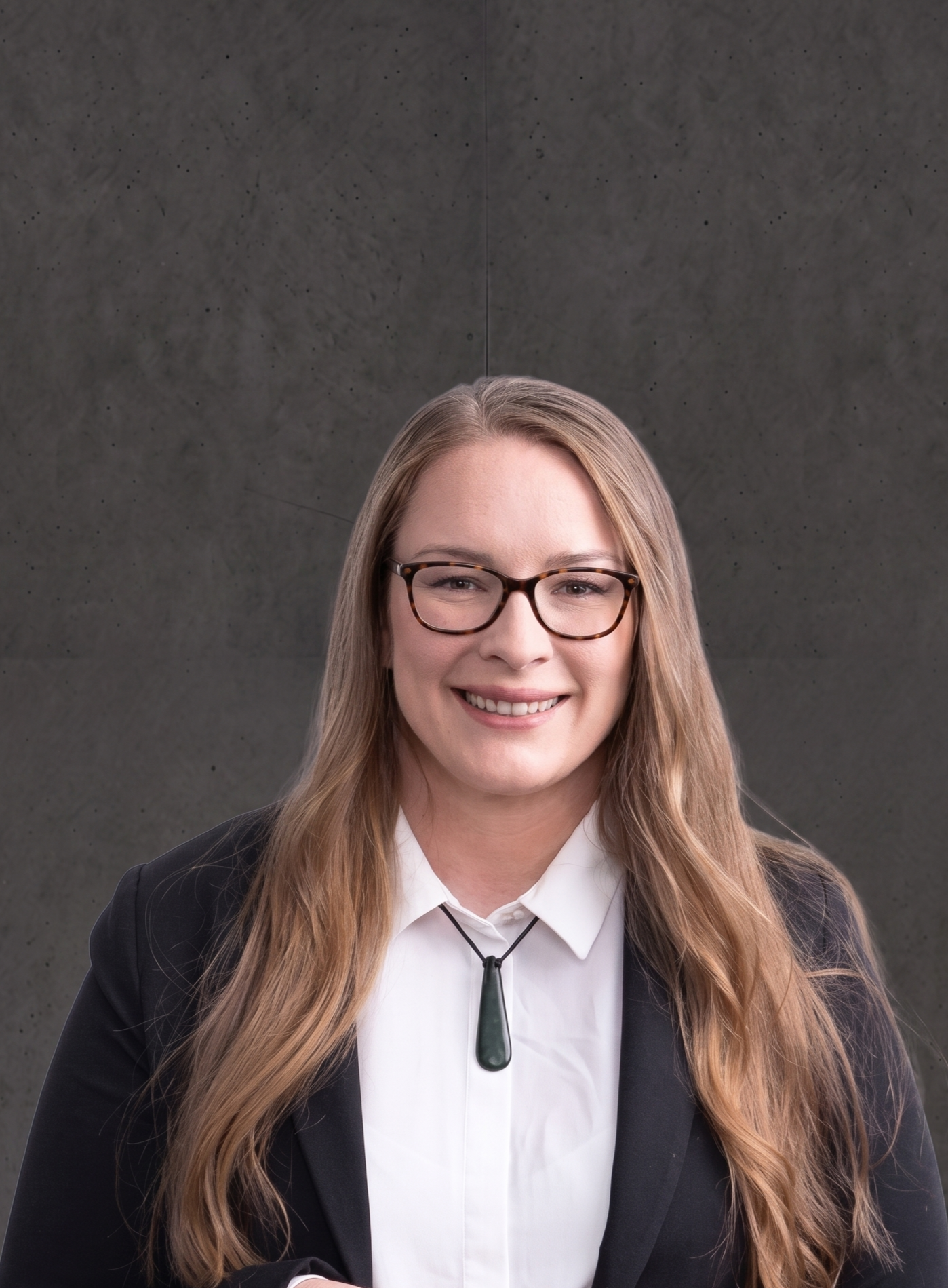 A young woman with long, wavy hair and glasses smiling, wearing a black blazer over a white shirt with a black tie and a hearing aid hanging around her neck, against a dark gray background.