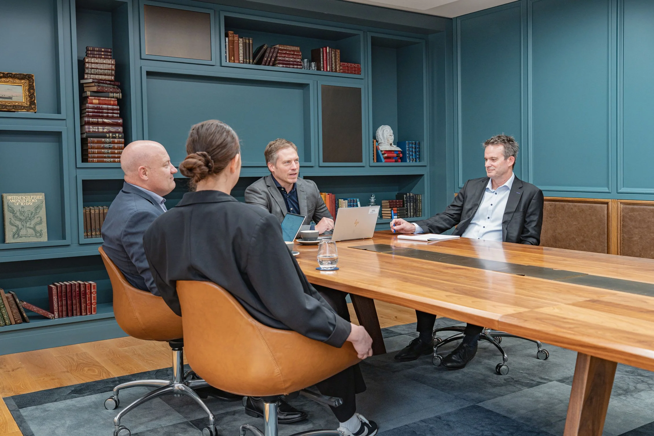 Four people seated around a wooden conference table, engaged in a discussion in a room with blue-green paneling and built-in bookshelves filled with books and decorative items.