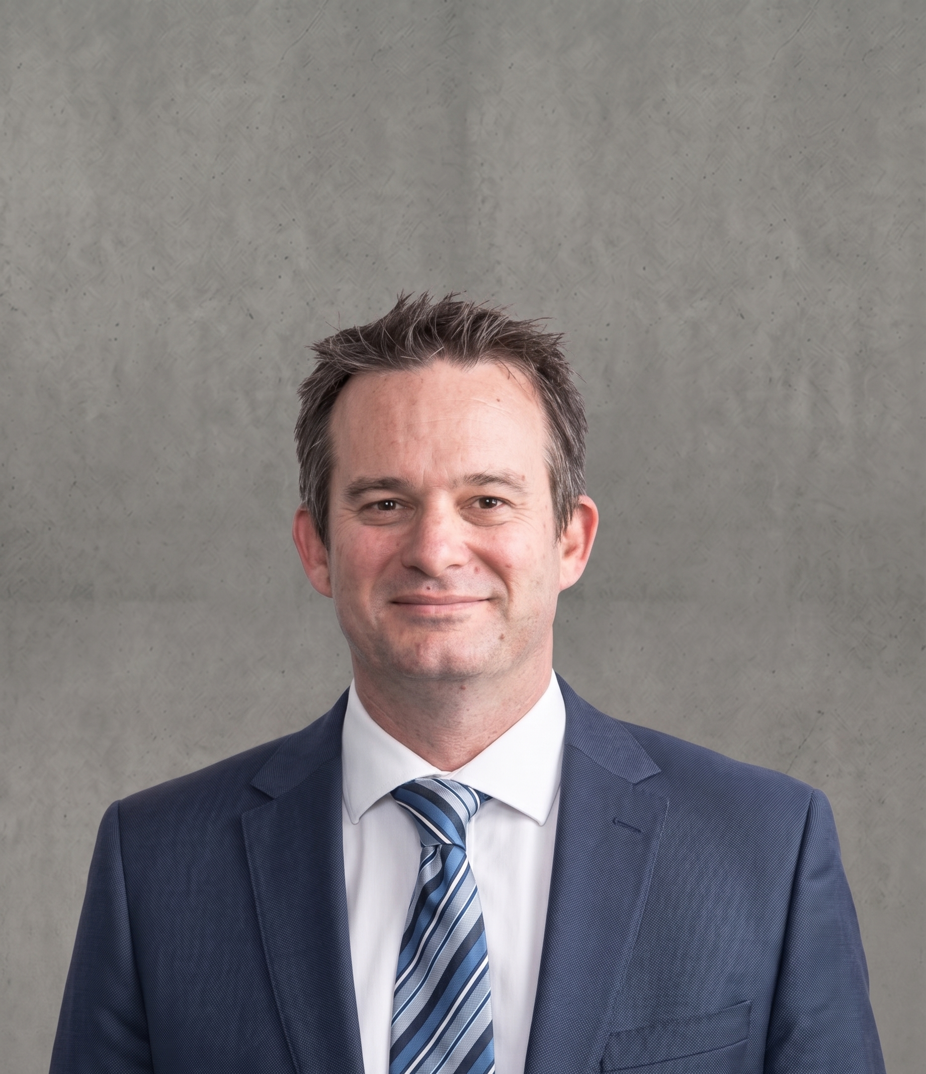 Close-up of a smiling man wearing a gray suit, white shirt, and striped blue and gray tie against a gray textured background.