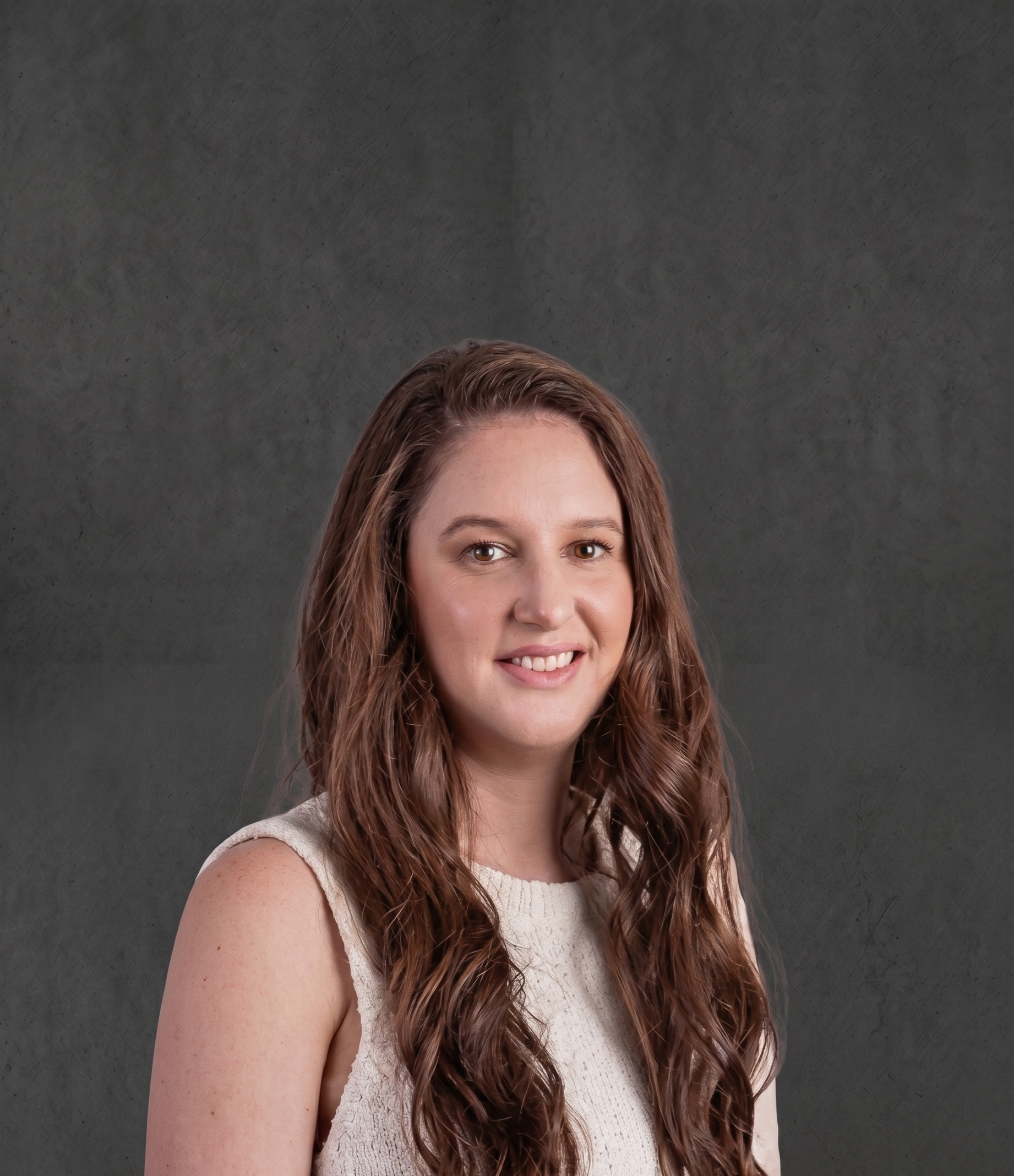 Close-up of a woman with long, wavy brown hair and brown eyes, wearing a sleeveless white lace top, smiling at the camera against a dark gray textured background.