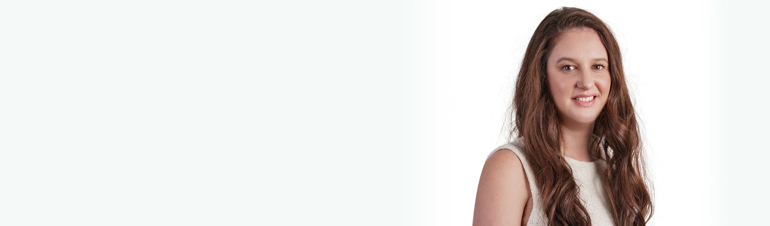 Portrait of a smiling woman with long wavy brown hair wearing a sleeveless beige top against a plain white background.