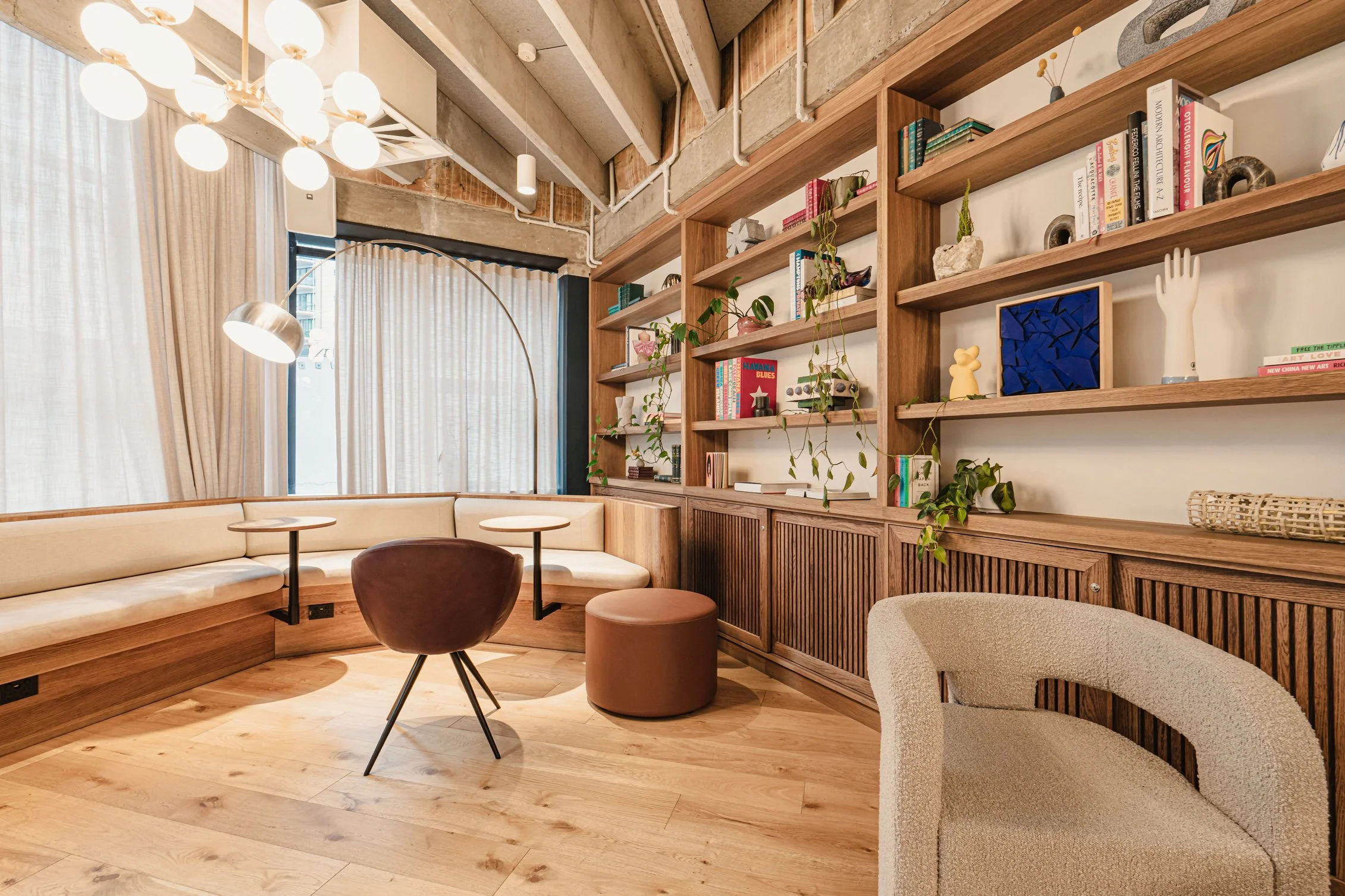 A cozy, modern living space with built-in wooden seating along the wall, featuring a beige cushion, small round tables, and a variety of books, plants, and decorative items on open wooden shelves. The room has large windows with curtains, a stylish ceiling light fixture, and a mix of wood and exposed brick and concrete ceiling elements.