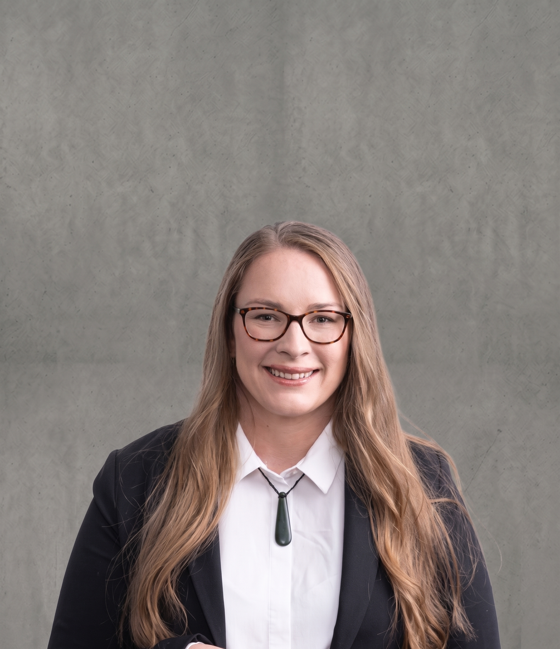 A smiling woman with long, wavy blonde hair, wearing glasses, a white collared shirt, and a black blazer, standing in front of a plain gray wall.