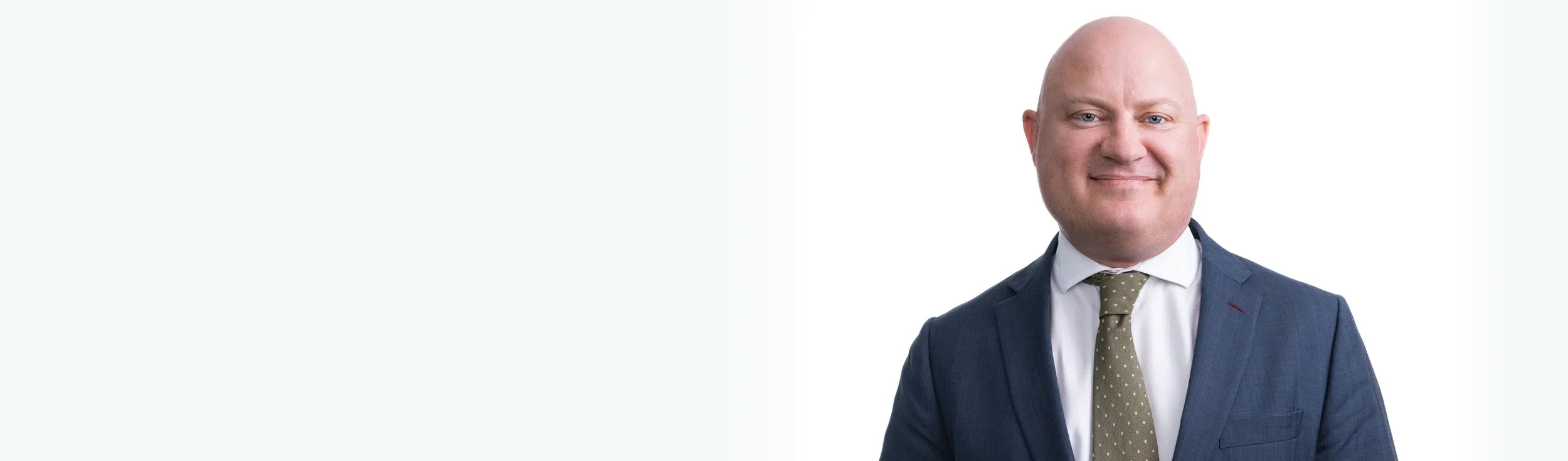 A professional man in a blue suit, white shirt, and polka dot tie smiling at the camera against a white background.