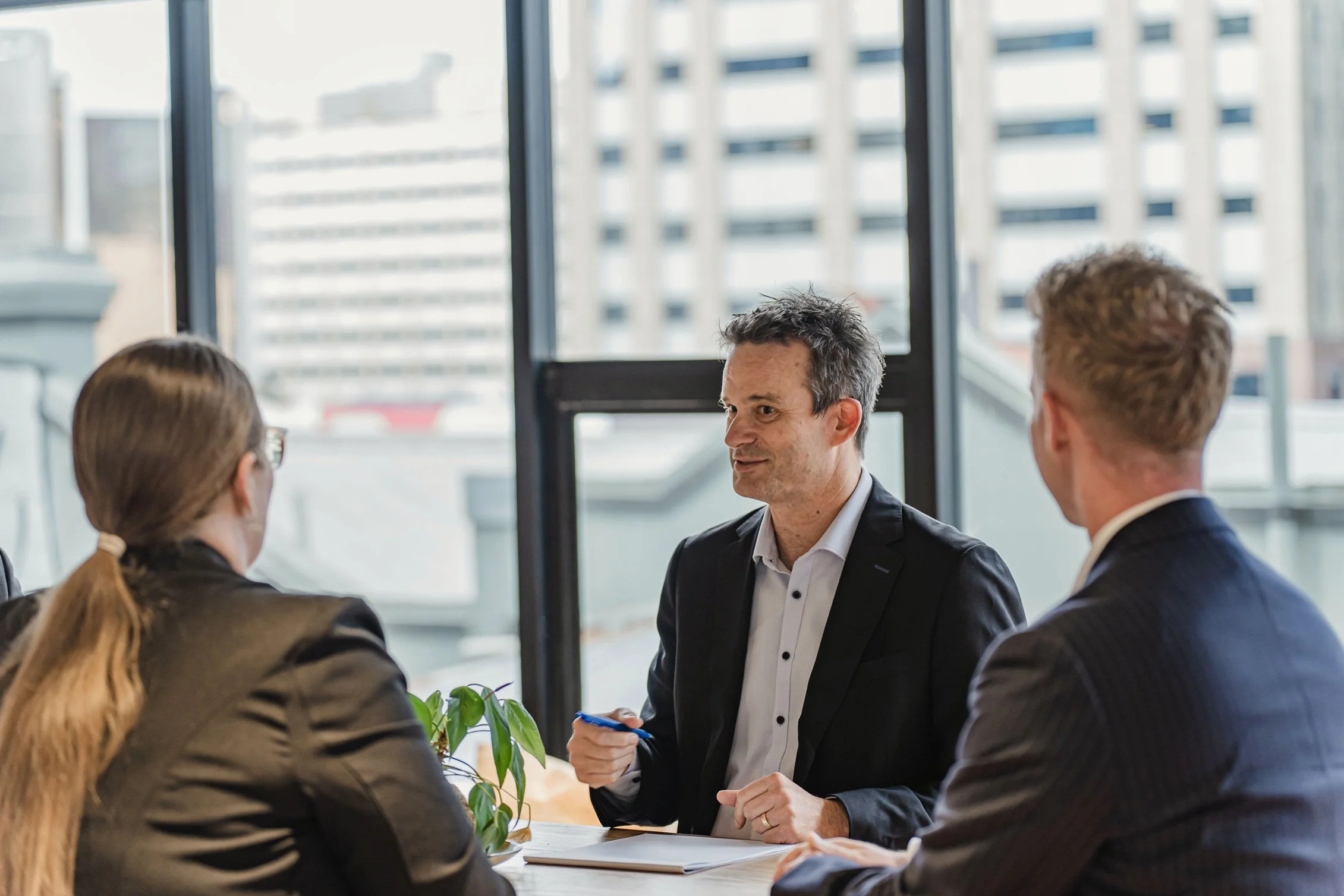 A group of four people having a discussion in a modern office with large windows and a cityscape view.