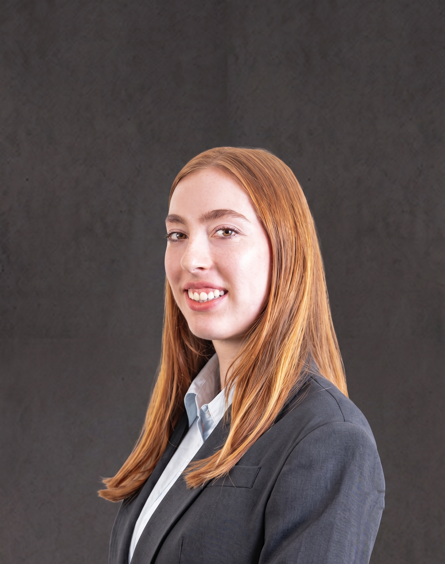 Close-up of a young woman with long, straight red hair, wearing a dark gray blazer and light blue shirt, smiling against a dark gray background.