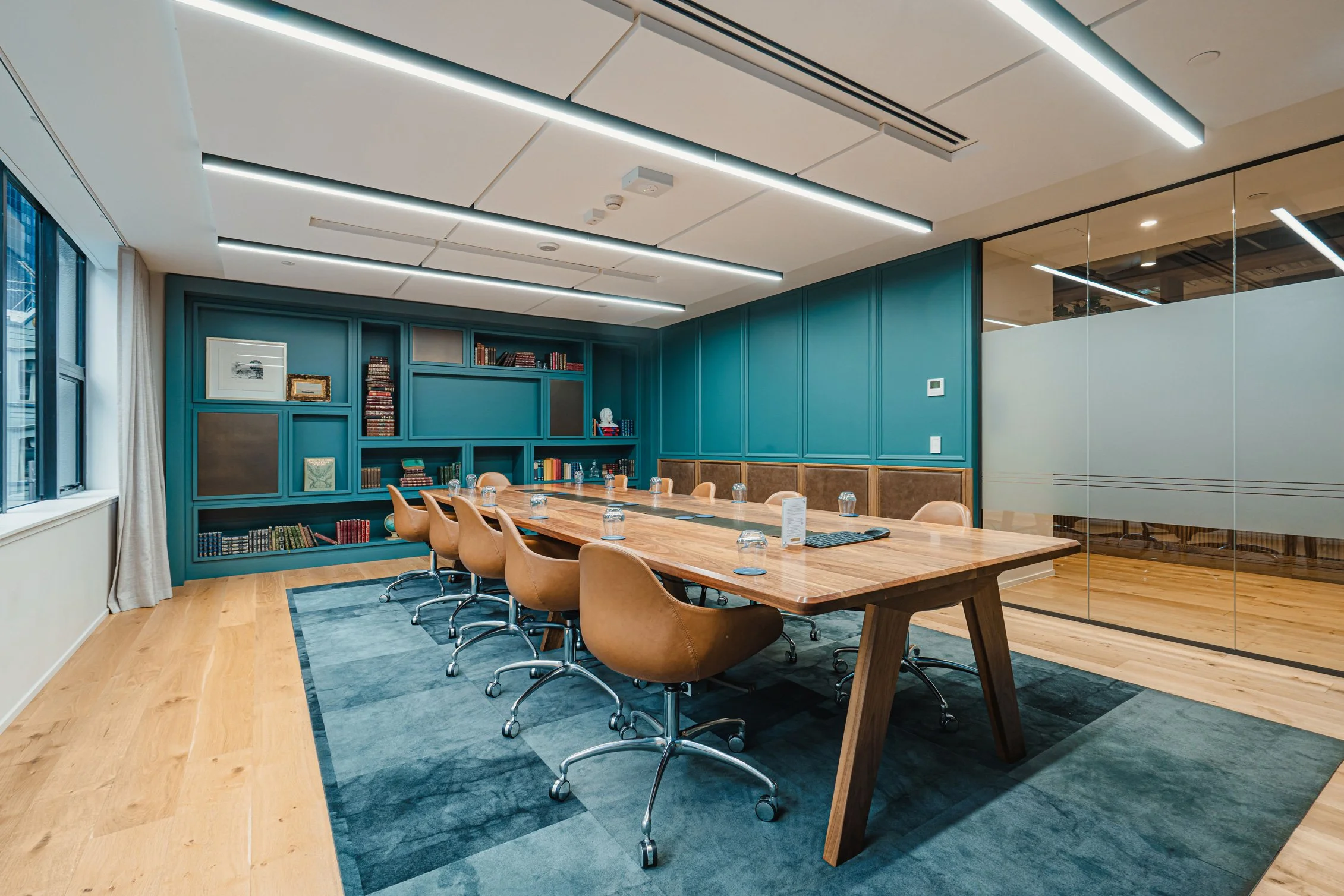 Modern conference room with a large wooden table surrounded by tan chairs. Blue paneled walls with bookshelves and framed artwork. Large windows with curtains and sleek ceiling lighting, hardwood floors, glass wall partition.