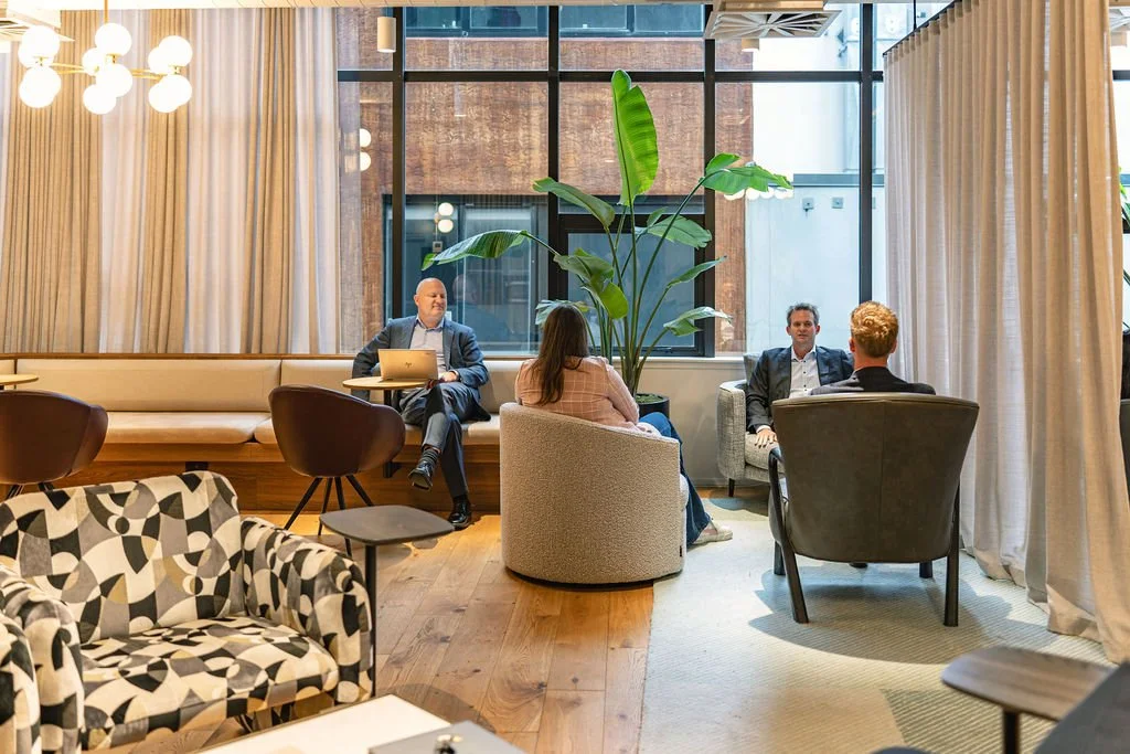 Four people sitting and having a discussion in a modern, well-lit lounge area with large windows, curtains, a green plant, and a mix of contemporary chairs and couches.