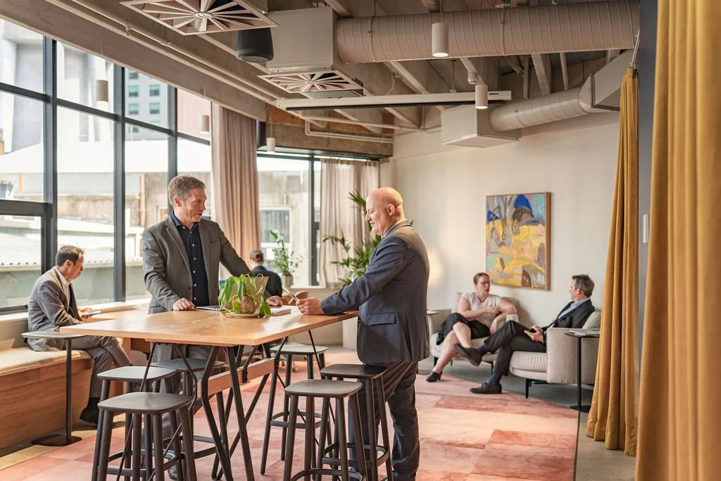 People in a modern office lounge area, with some sitting and others standing, engaging in conversation. The room has large windows, art on the wall, and gold curtains.