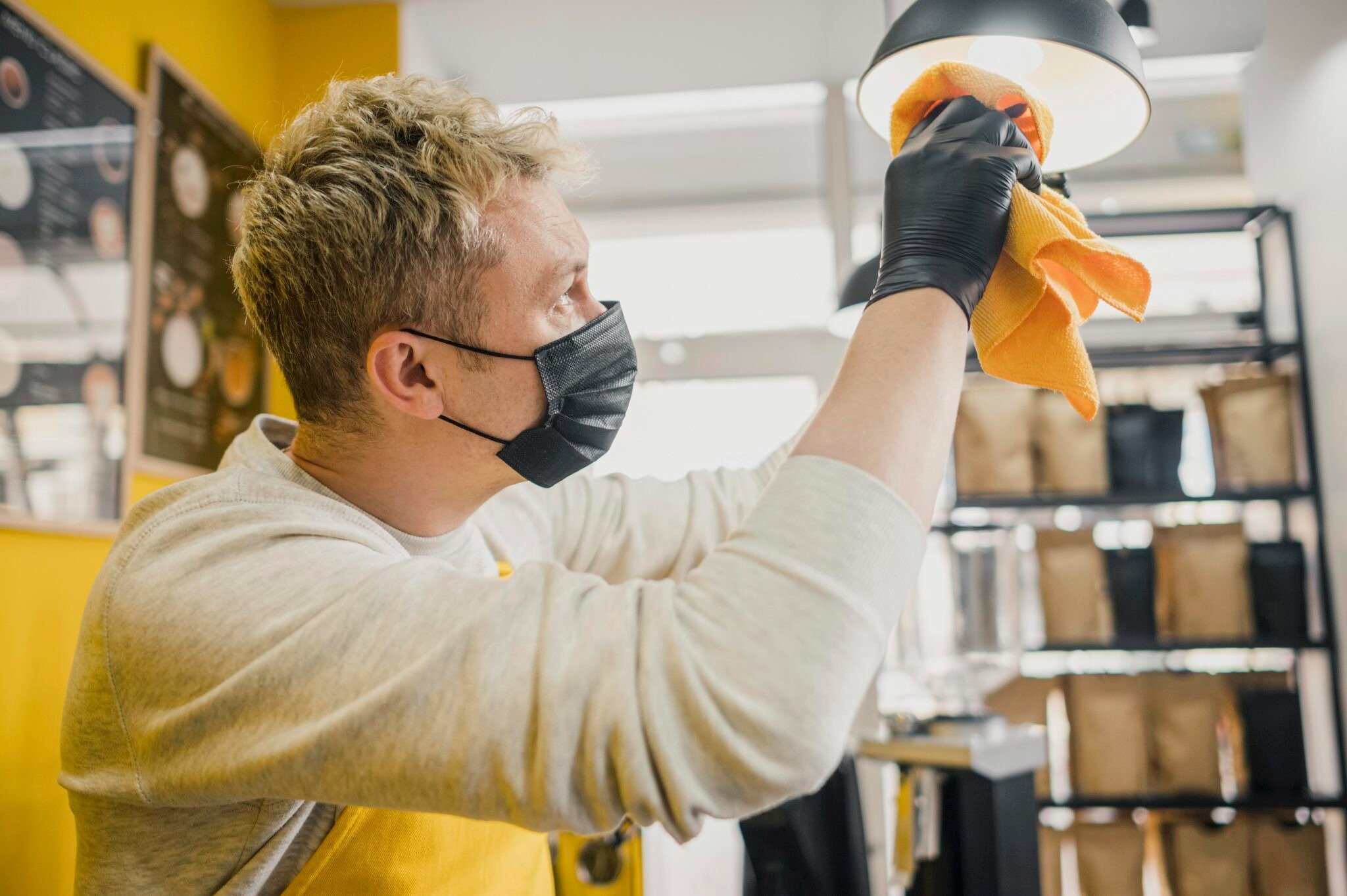 A person wearing a black face mask and black gloves cleaning a light fixture with an orange cloth in a cafe or restaurant setting.