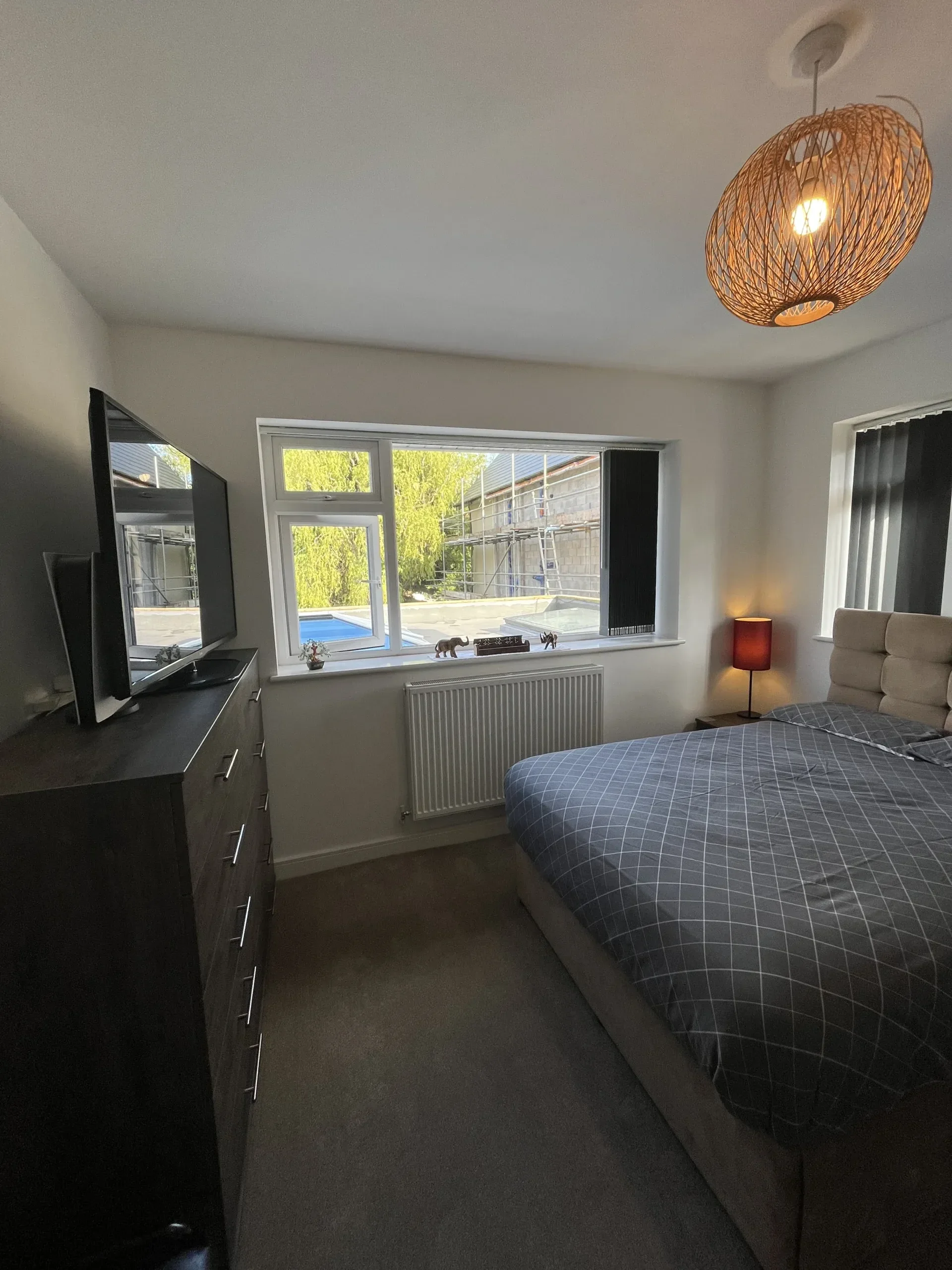 A bedroom with a large window showing a garden with trees outside. The room has a bed with a checkered blanket, a bedside lamp, and a dresser with a TV on top. There is a ceiling light fixture made of woven material.
