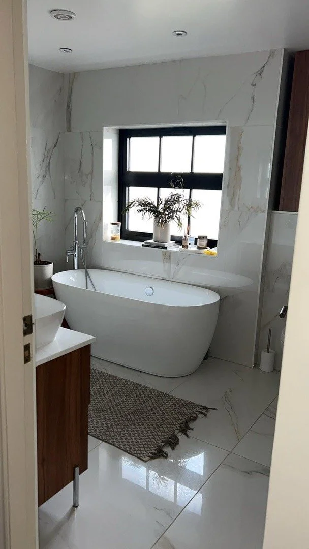 A modern bathroom with a white bathtub beneath a window, gray marble walls, a plant on the window sill, a wooden vanity with a vessel sink, and a woven rug on a shiny marble floor.