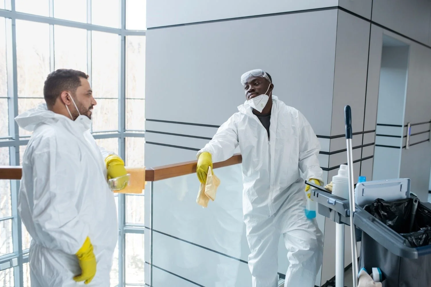 Two healthcare workers in white protective suits, face masks, and yellow gloves, engaging in a conversation in a modern building corridor, with cleaning equipment nearby.