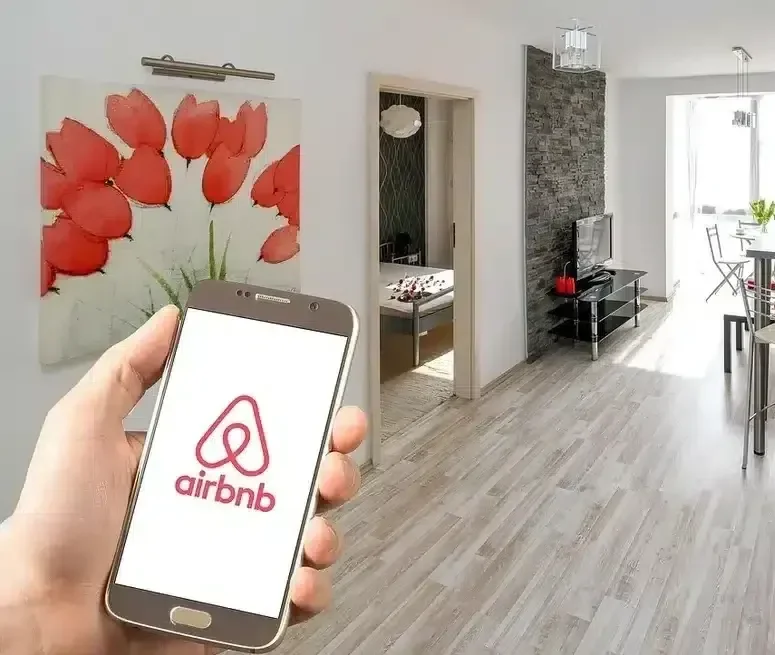 A person holding a smartphone displaying the Airbnb logo in a modern apartment living room with a large floral painting, a black stone accent wall, a TV on a stand, and a dining area with chairs and a table.