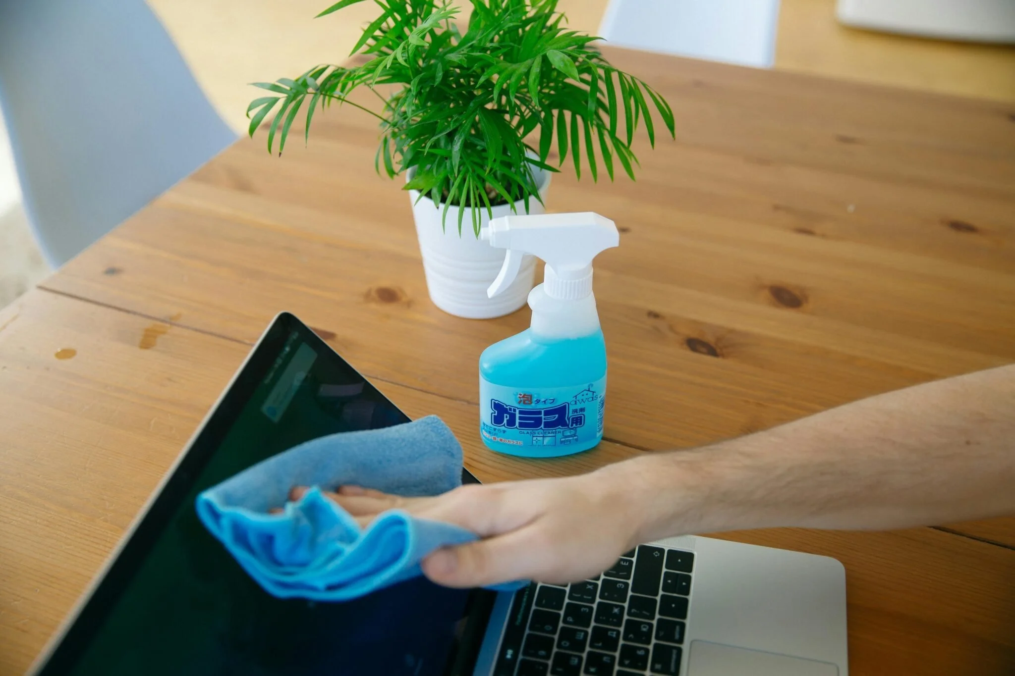 A person is cleaning a laptop screen with a blue cloth, balancing their arm on the table. There is a spray bottle with blue cleaning liquid and Japanese writing on the table, next to a potted green plant on a wooden table.