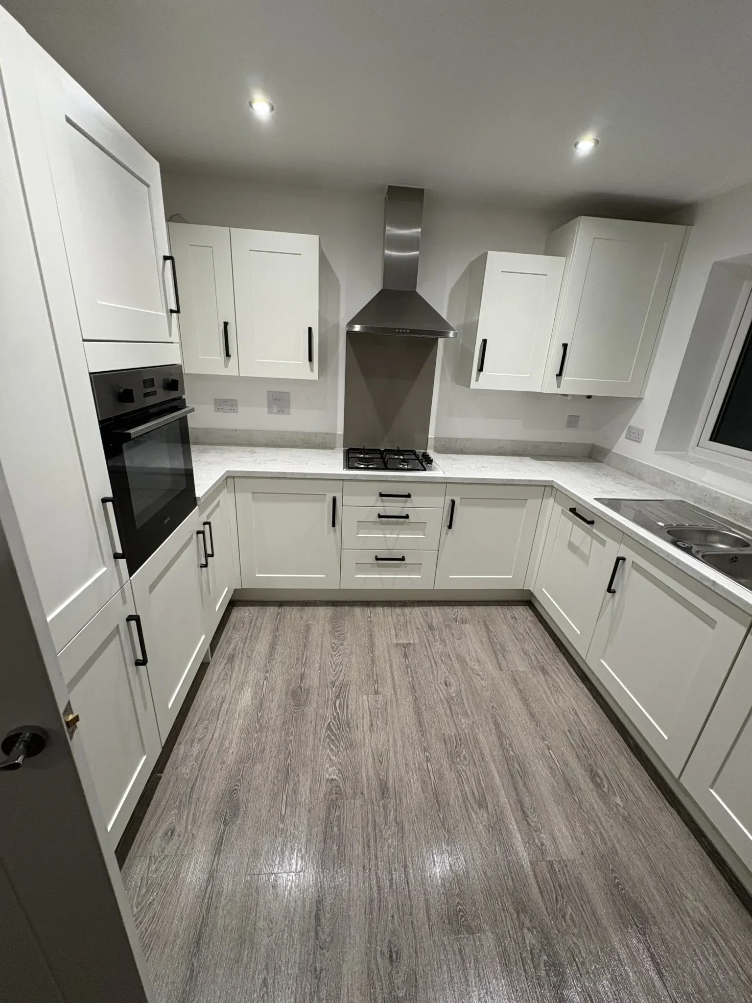 Modern kitchen with white cabinets, a stainless steel oven, a gas stove, a range hood, and a double sink, with wood-look flooring and recessed ceiling lights.