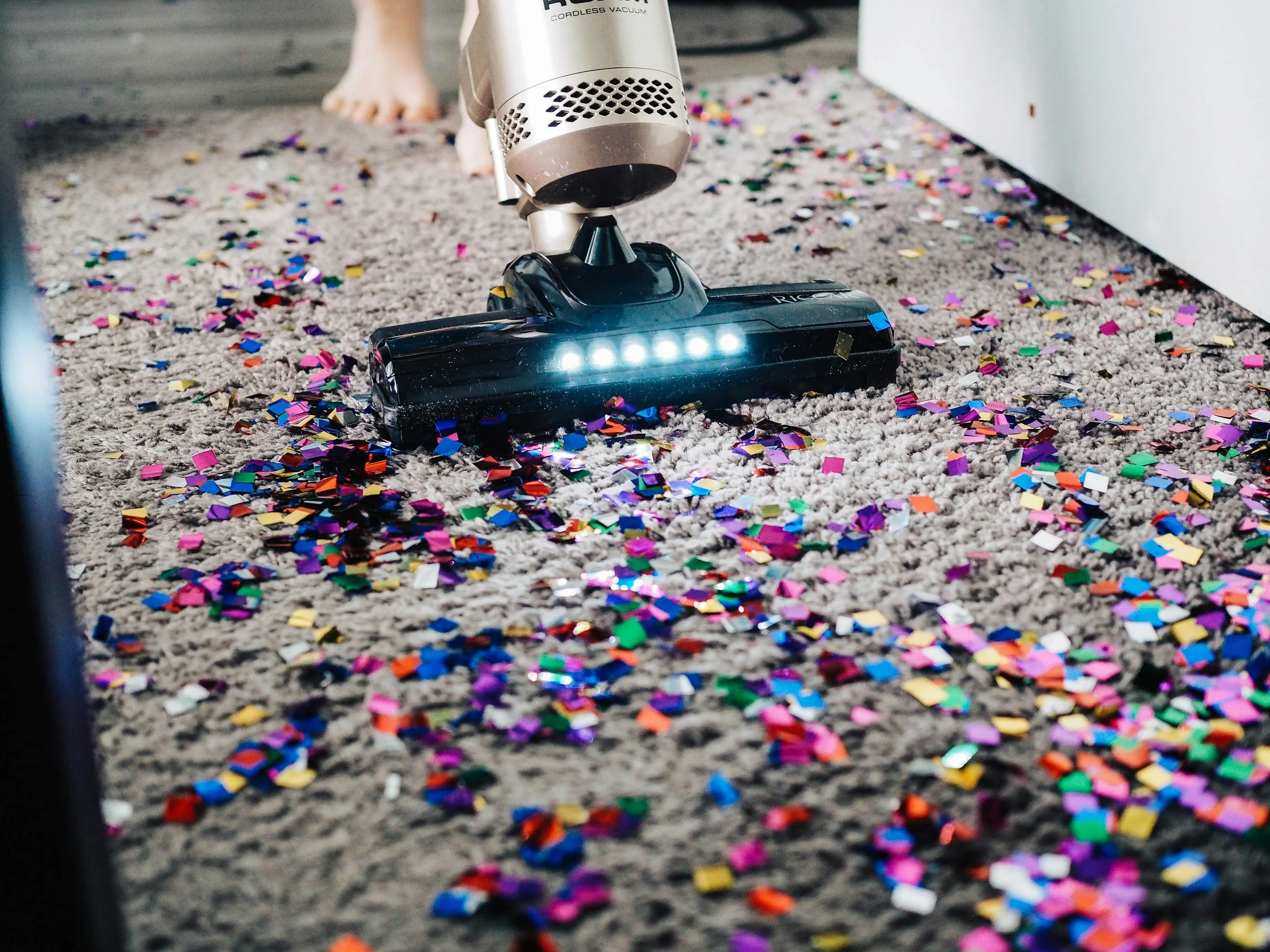 Vacuum cleaner cleaning up colorful confetti on a gray carpet with a person's foot visible in the background.