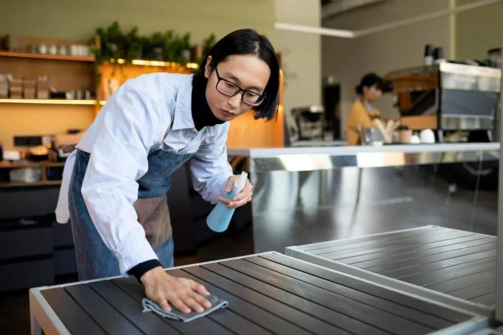 A person wearing safety glasses and a gray apron disinfects a restaurant table with a spray bottle and cloth.