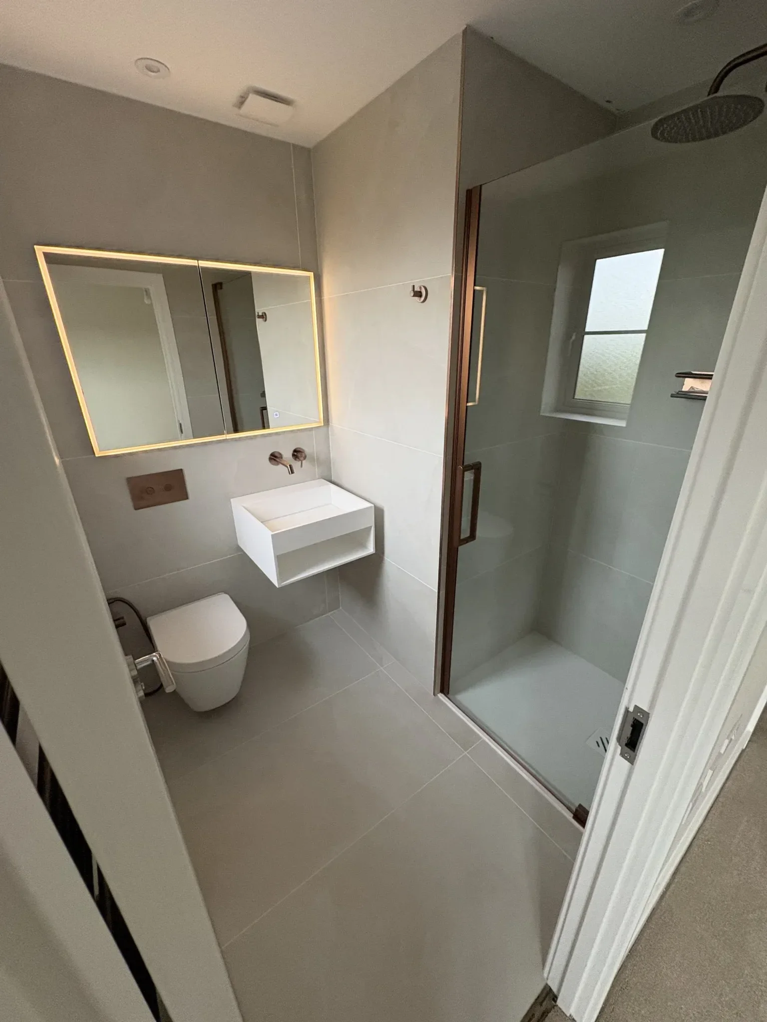Modern minimalist bathroom with a wall-mounted toilet, small sink, large mirror with backlighting, walk-in shower with a glass door, gray tile flooring and walls, and a small window.