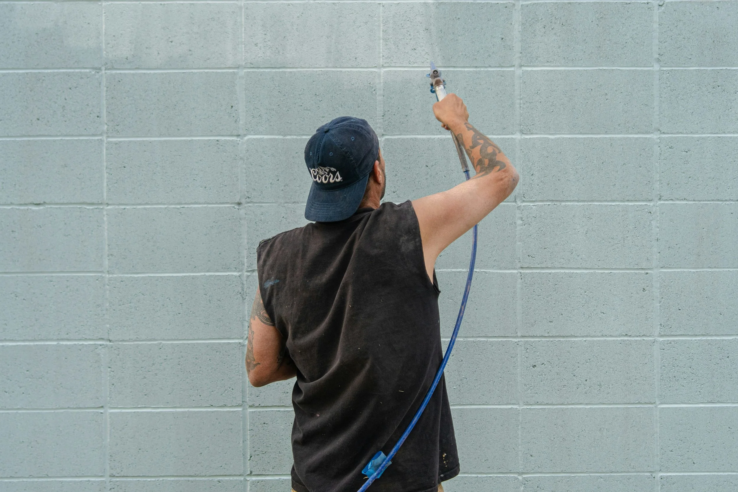 A person with tattoos on their arm, wearing a black sleeveless shirt and a blue cap, is pressure washing a light blue cinder block wall.