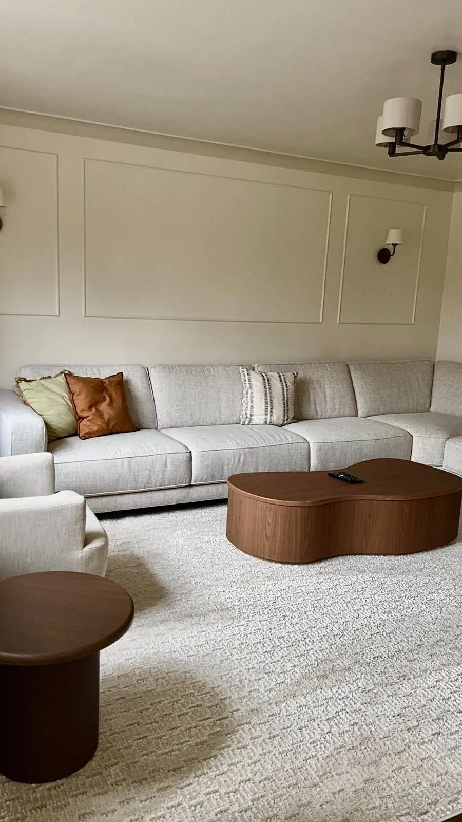 Living room with a long beige sectional sofa, decorative pillows, a wooden coffee table, and wall-mounted light fixtures.