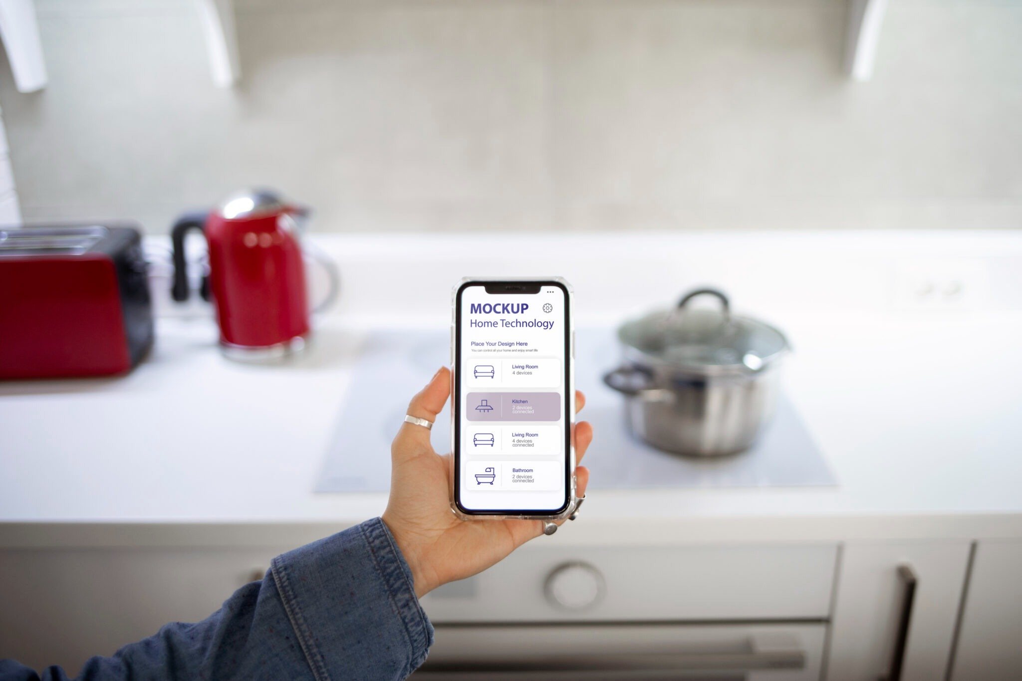Person holding a smartphone displaying a home automation app in a kitchen, with a stovetop and kitchen appliances in the background.