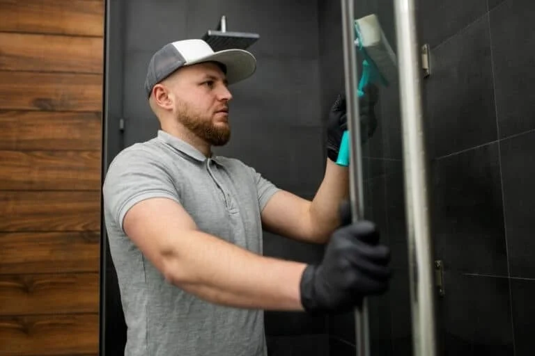 A man wearing a gray cap, gray polo shirt, and black gloves is cleaning or inspecting the inside of a metal locker or cabinet in a room with dark walls and wood paneling.