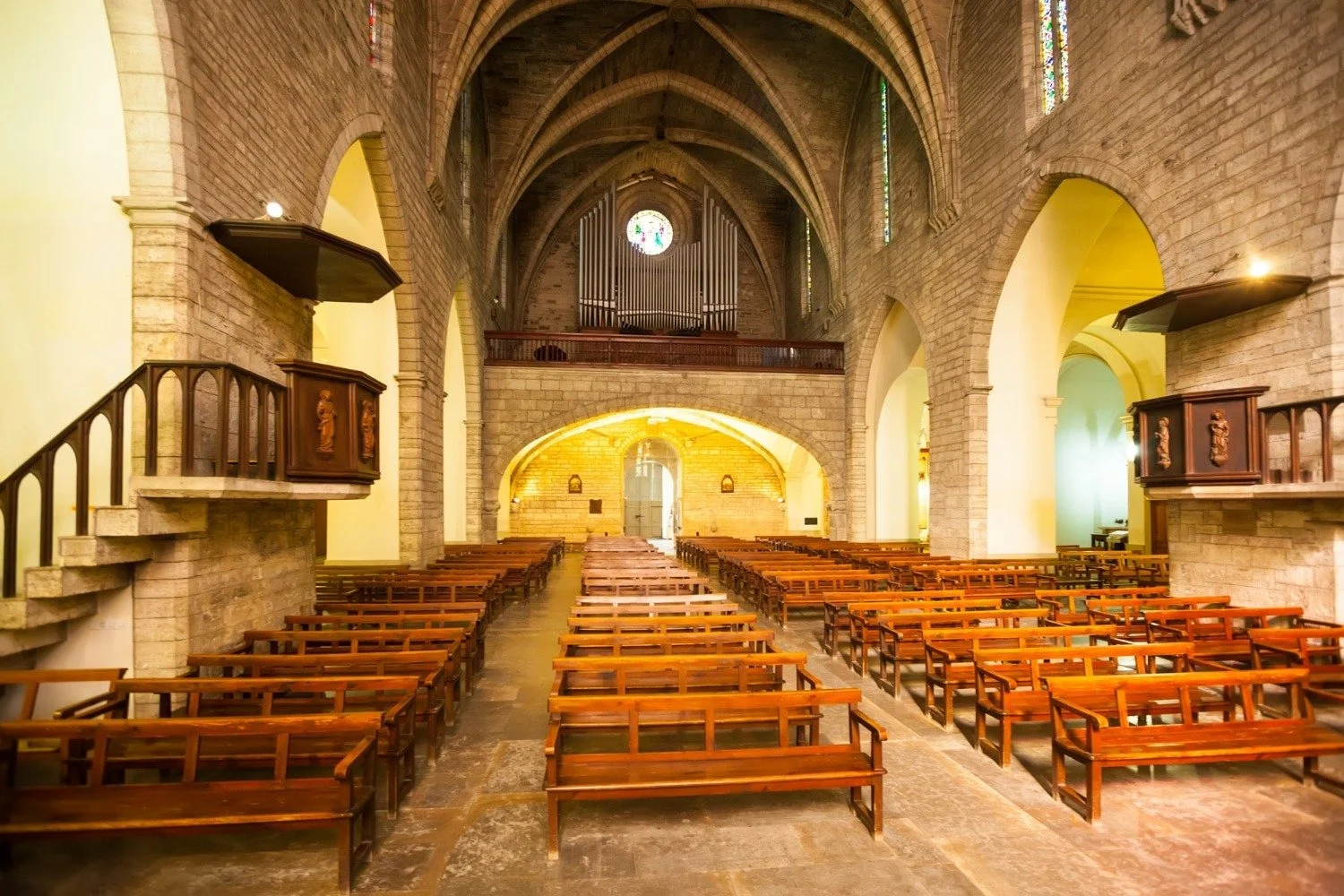 Interior of a church with wooden pews, stone walls, high vaulted ceilings, stained glass windows, and an organ at the back.