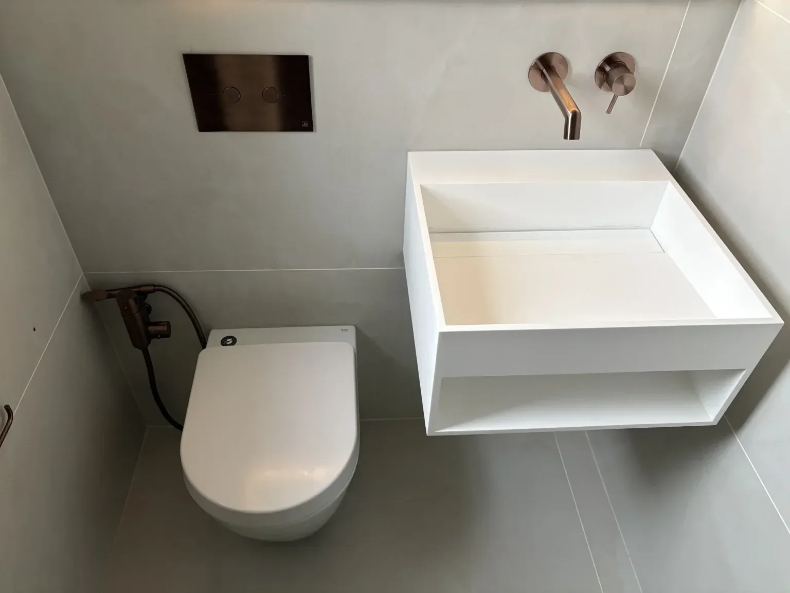 Modern bathroom with a white toilet and a white sink with a brass faucet. The wall behind has light-colored tiles, a stainless steel flush panel, and a brass water outlet.