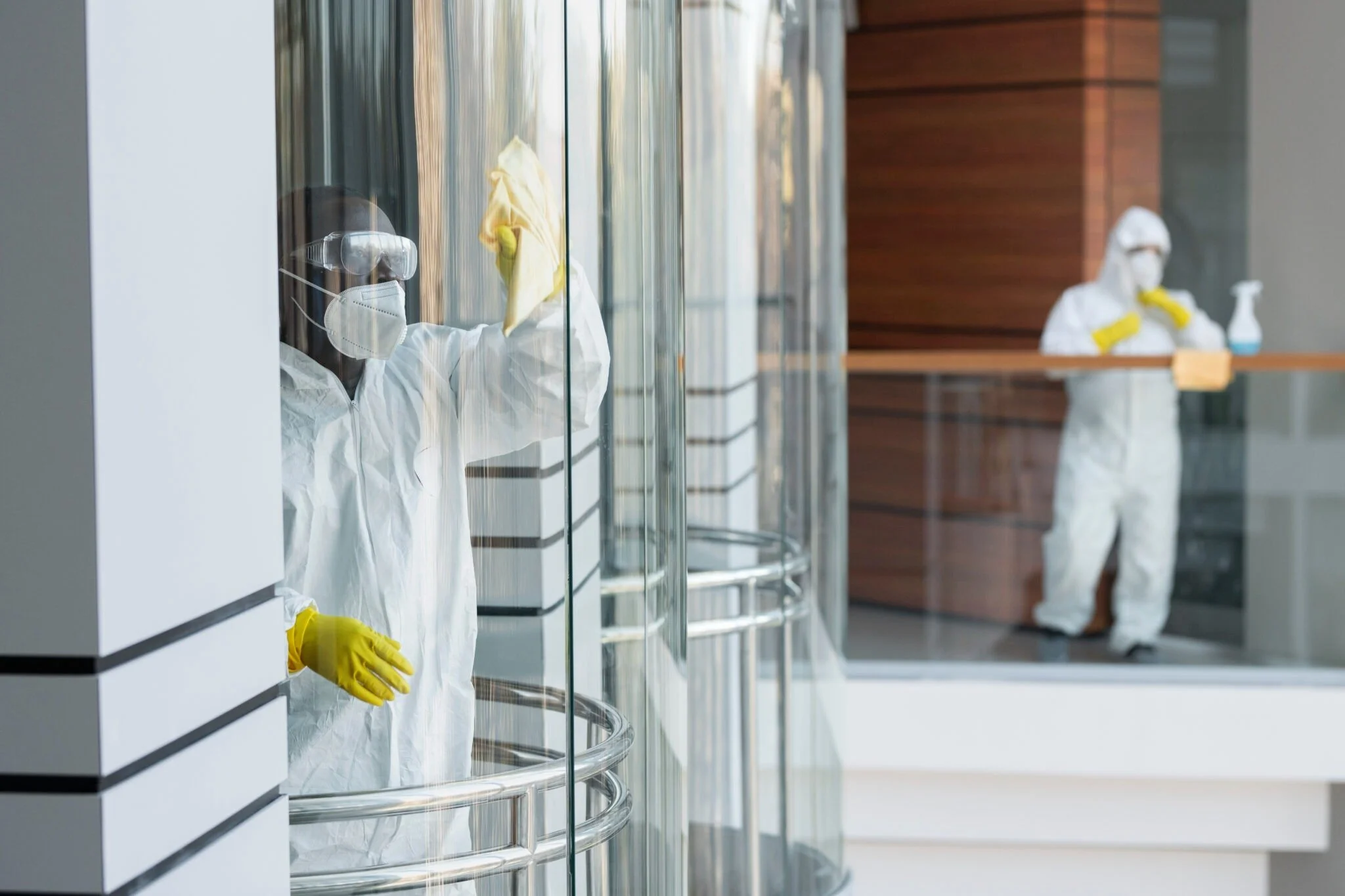 Two healthcare workers in full protective gear cleaning glass windows, one in the foreground wiping with a cloth and another in the background standing at a counter with cleaning spray.