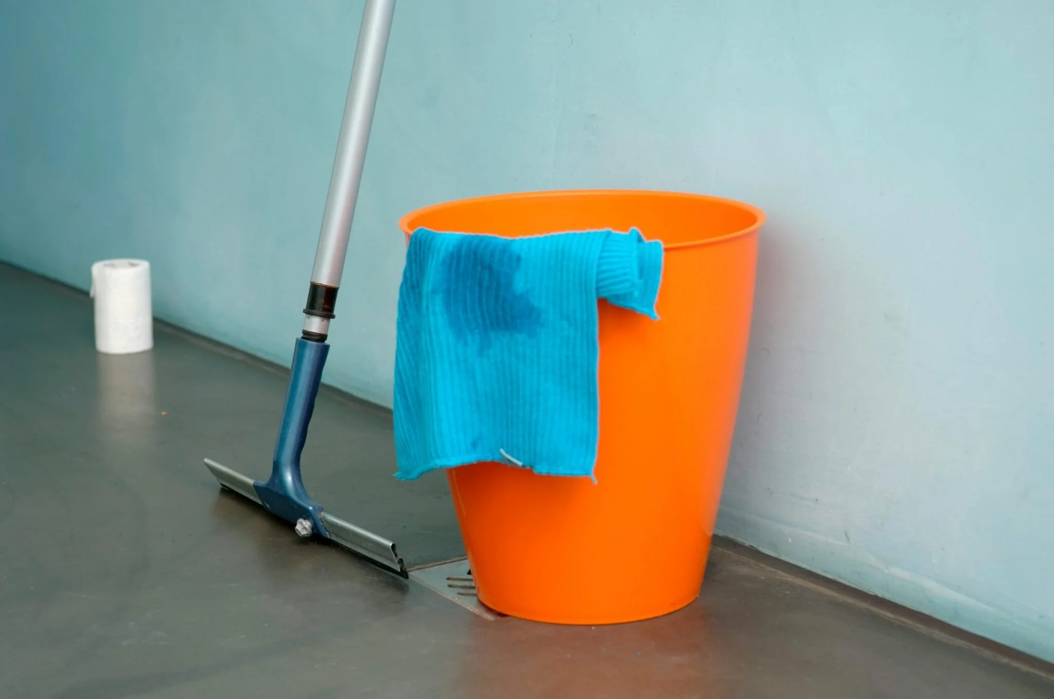 An orange bucket with a blue cleaning cloth draped over its edge, placed on a floor next to a mop and a paper towel roll, against a light blue wall.