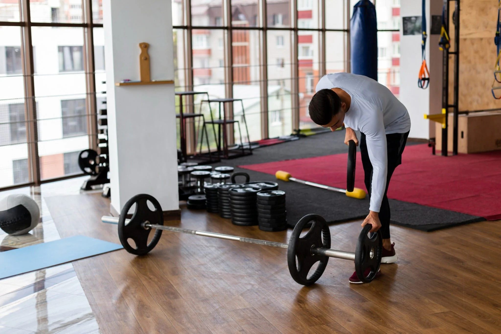 A man is preparing to lift a barbell with weights in a gym. The gym has large windows, various weights, mats, and exercise equipment in the background.