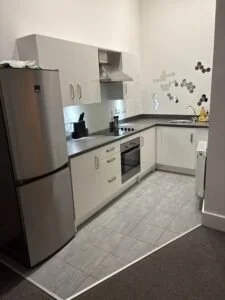 Kitchen with white cabinets, gray countertops, a refrigerator, a stove, and a microwave, with some wall decor and a small trash can.