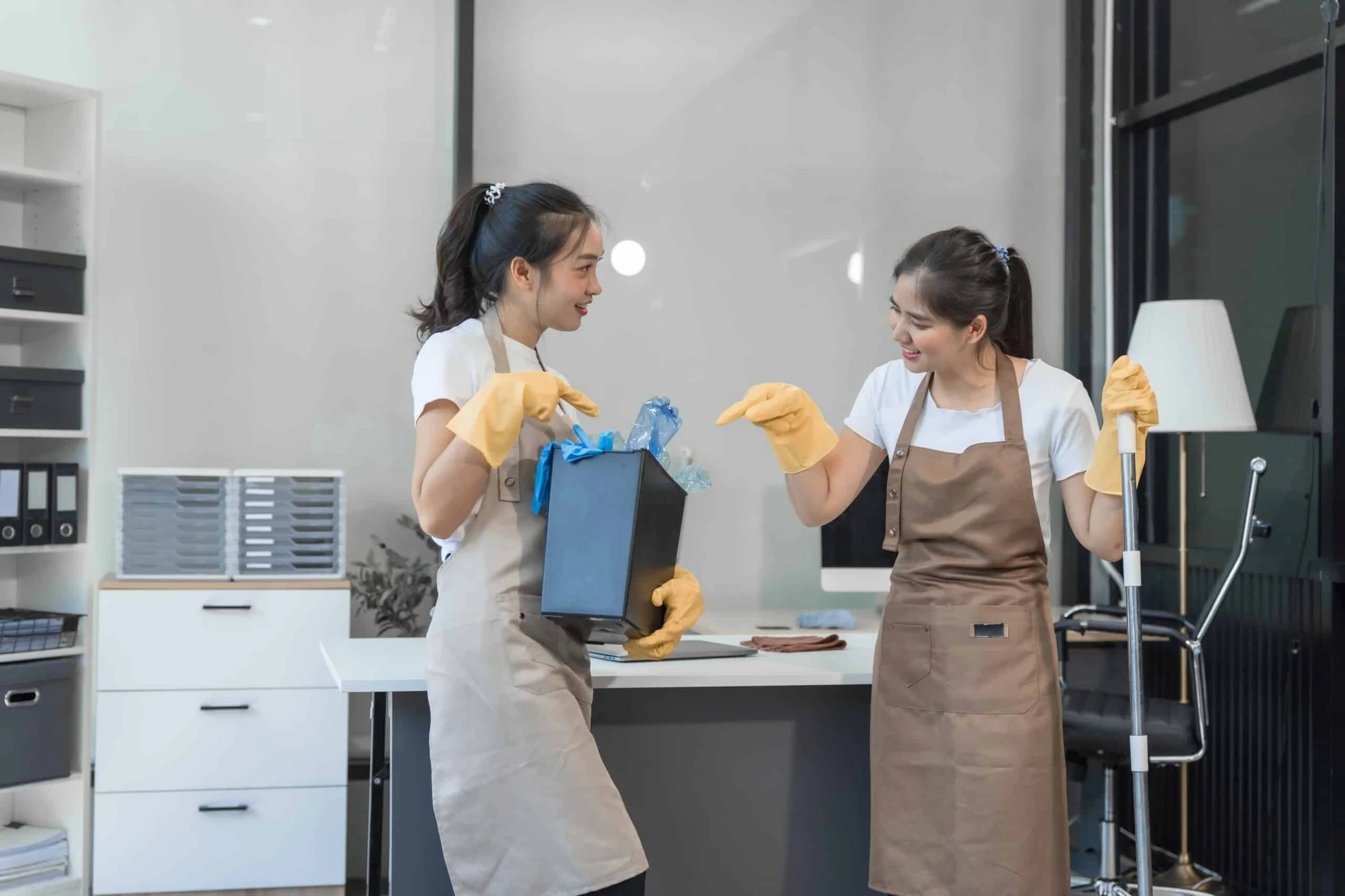 Two women wearing aprons and yellow gloves laughing and joking in a cleaning or office workspace.