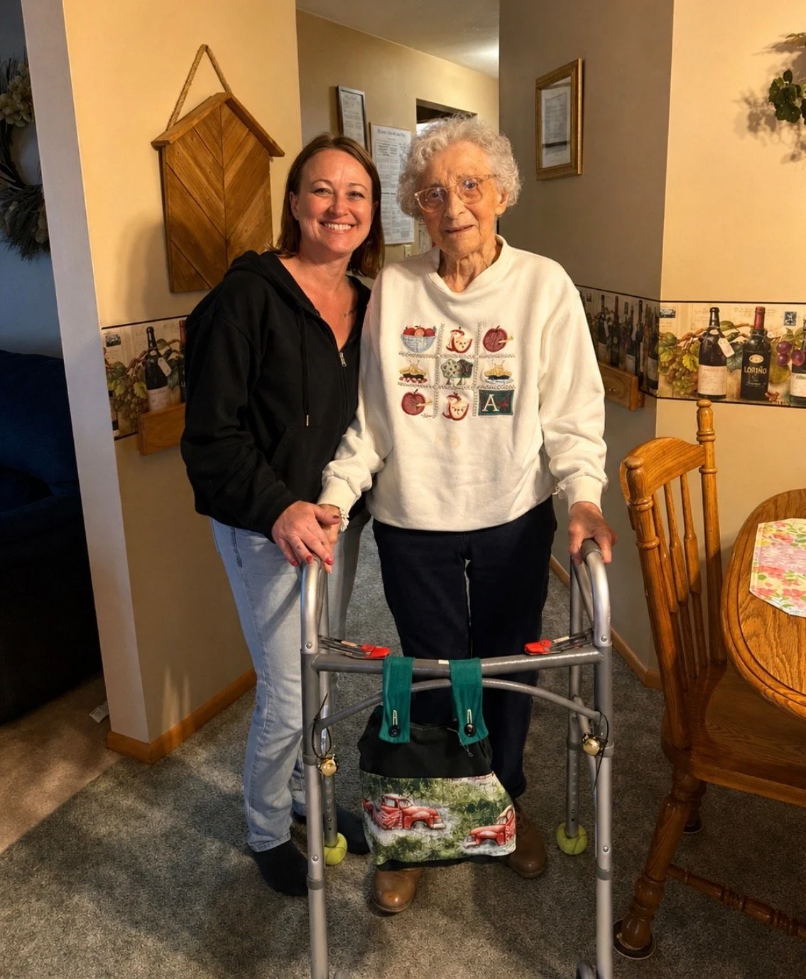 A young woman and an elderly woman standing together indoors, smiling. The elderly woman is using a walker and wearing a sweatshirt with colorful embroidered designs. The young woman is holding the elderly woman's hand, and there are dining chairs an