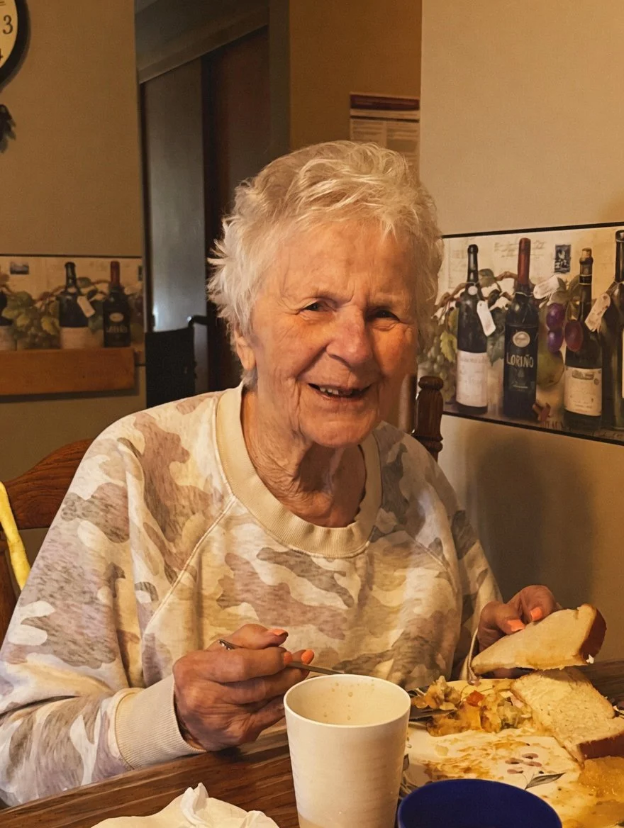 An elderly woman with short gray hair smiling and sitting at a table. She is holding a slice of bread and has a plate with food in front of her. The background shows a wall with pictures of wine bottles.