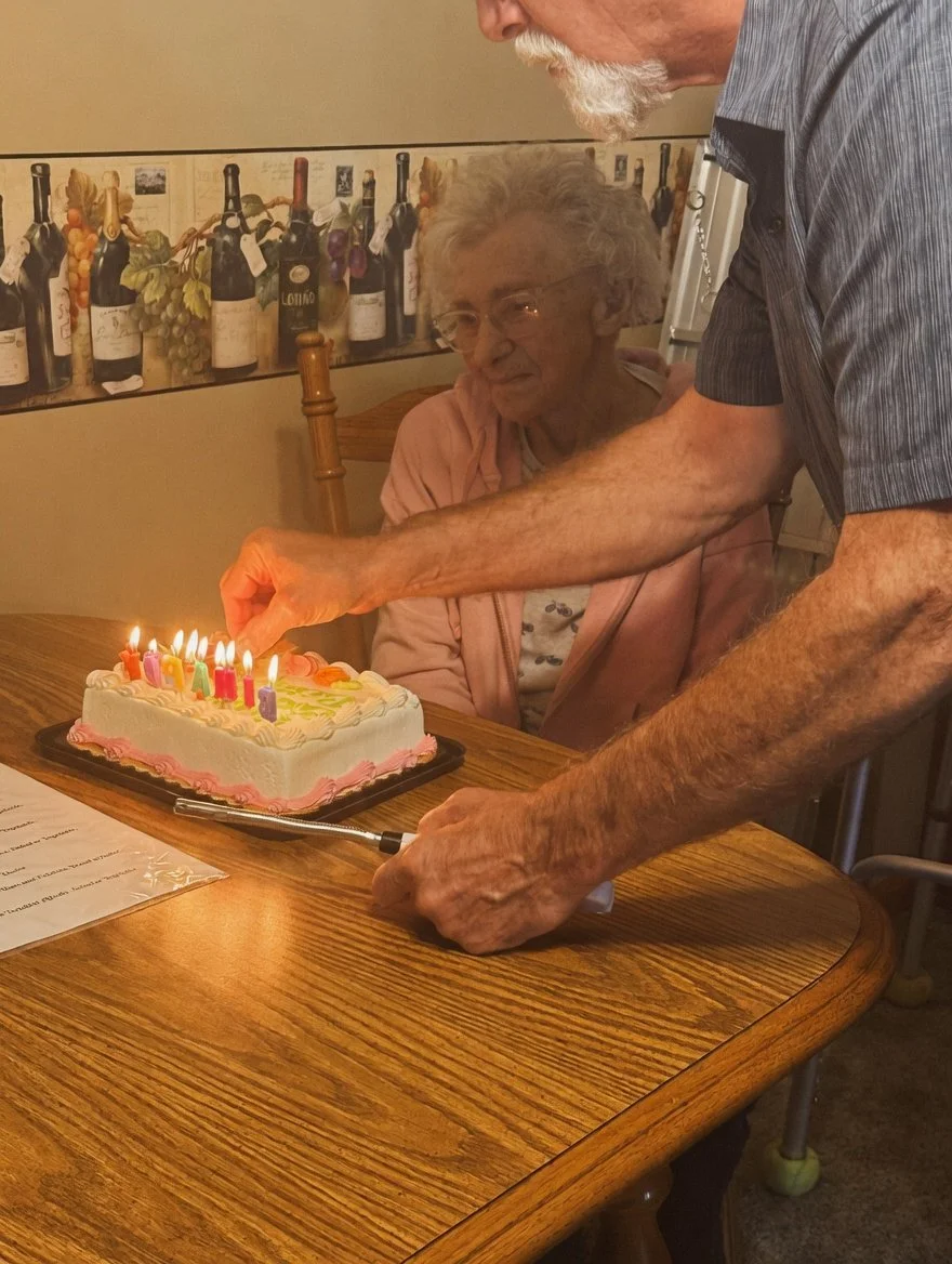 An elderly woman with glasses and a pink jacket is sitting at a wooden table, watching a man light birthday candles on a cake with pink and white frosting. The setting appears to be in a dining area with a decorative wine bottle and grapevine-themed 
